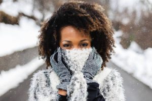 Woman covering lower half of face with mittens and scarf snowy background